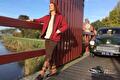 Vrouw in rode jas en laarzen leunt op brug, oldtimer erachter