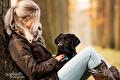 Blonde vrouw in waxjas met zwarte labrador tegen een boom